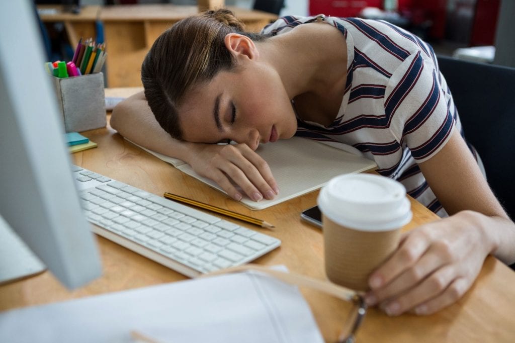 Student slumped over desk sleeping with coffee in hand